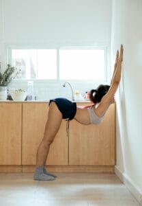 Full body side view of anonymous active female in sportswear practicing exercise with hands on wall and bending back in kitchen