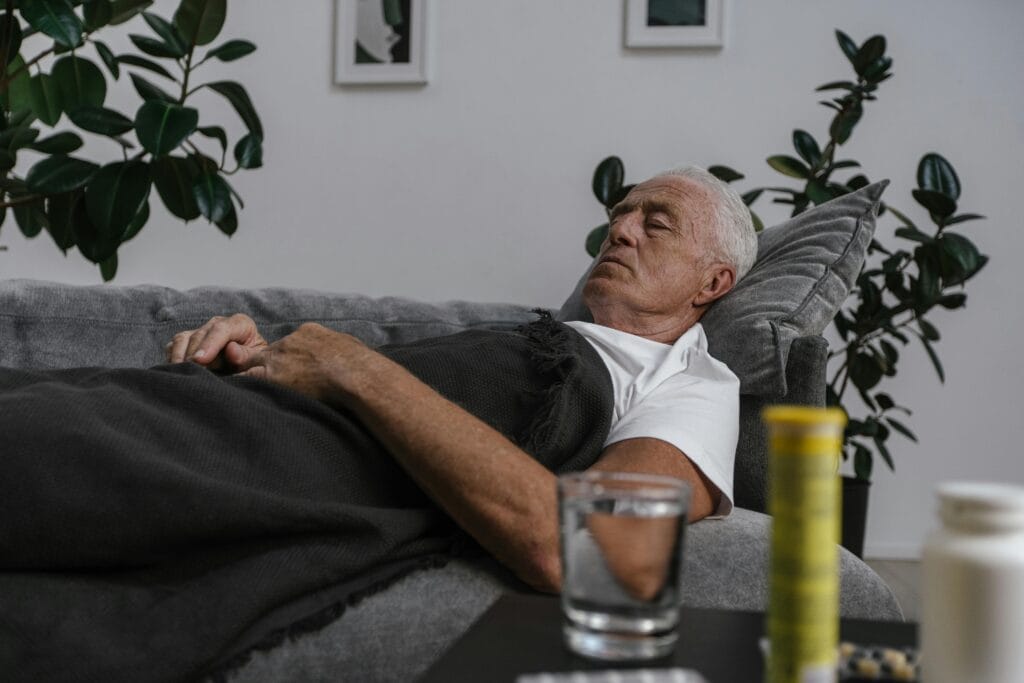 An elderly man sleeping on a couch with a blanket and glass of water nearby.