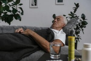 An elderly man sleeping on a couch with a blanket and glass of water nearby.