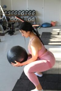 A woman in sportswear performs squats with a medicine ball indoors, emphasizing fitness and strength.