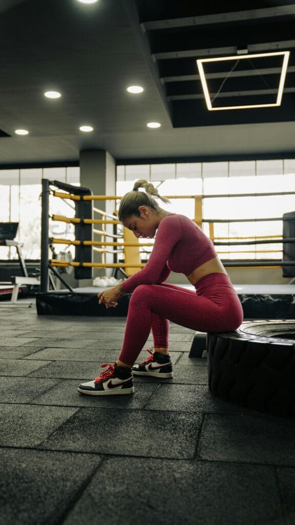 Female athlete rests in a gym near a boxing ring, wearing vibrant sportswear.