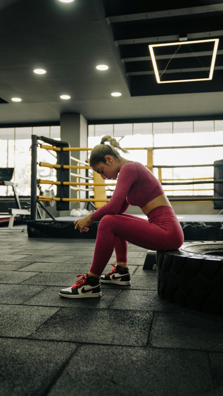 Female athlete rests in a gym near a boxing ring, wearing vibrant sportswear.