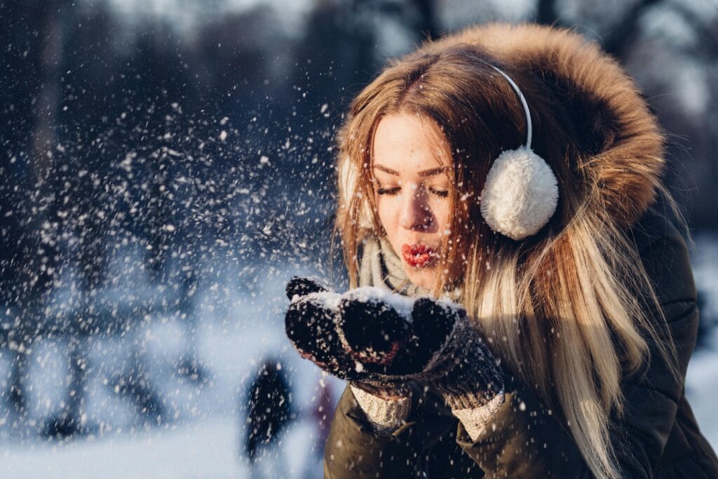 A young woman enjoying a snowy day, blowing snow while wearing cozy earmuffs and a winter coat.