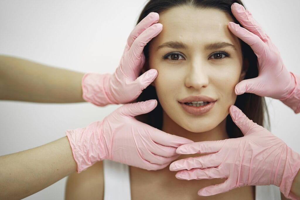 A woman receiving a facial treatment, hands in gloves gently touching her face indoors.