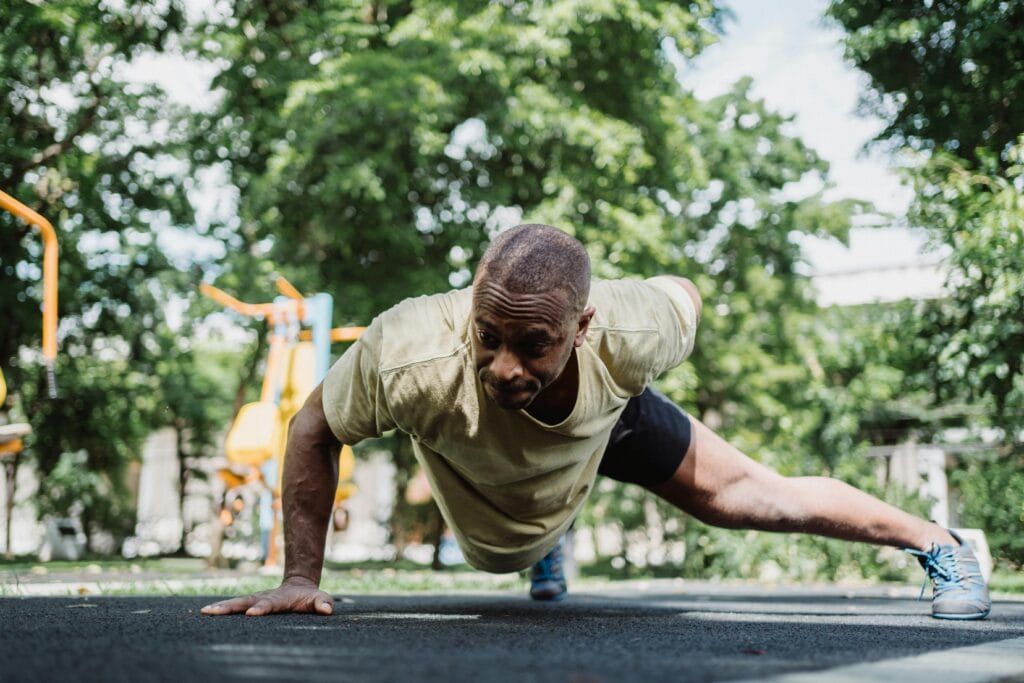 Athletic man performs a one-arm push-up in a lush park setting, showcasing strength and fitness.