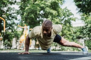 Athletic man performs a one-arm push-up in a lush park setting, showcasing strength and fitness.