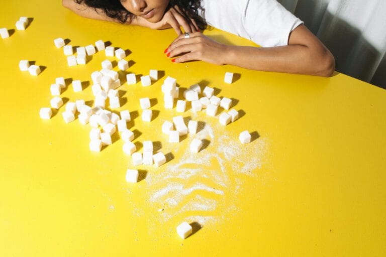 A woman contemplating sugar cubes on a bright yellow table, symbolizing health choices.