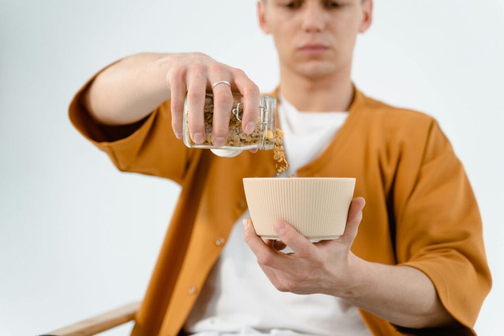 A man in a brown shirt pours cereal from a glass jar into a bowl, focused on the task.