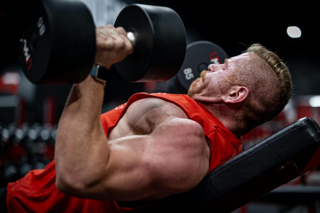 Side view of a muscular man lifting dumbbells on a bench in a gym, focusing on strength and effort.