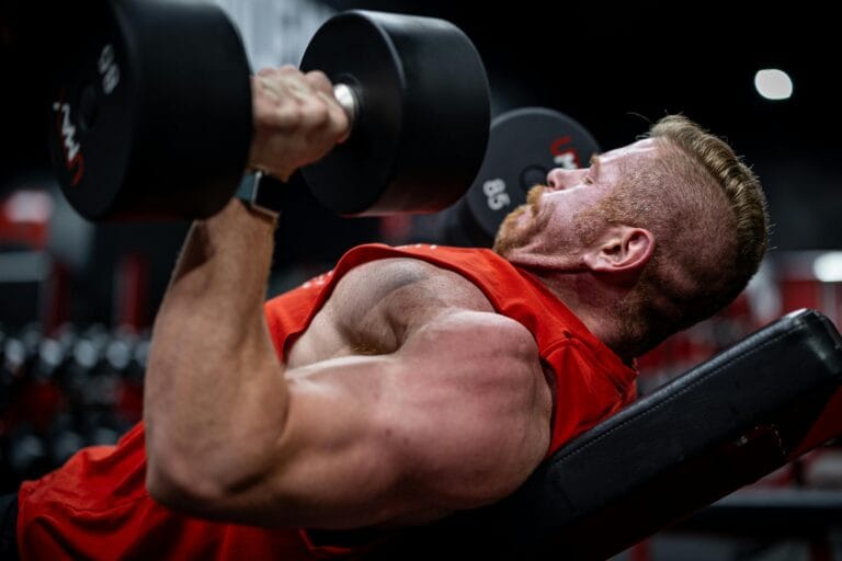 Side view of a muscular man lifting dumbbells on a bench in a gym, focusing on strength and effort.