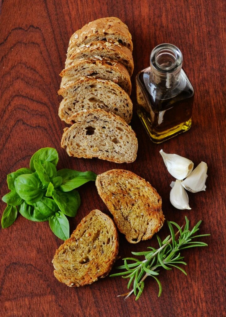 Sliced rustic bread with olive oil, basil, rosemary, and garlic on a wooden table.