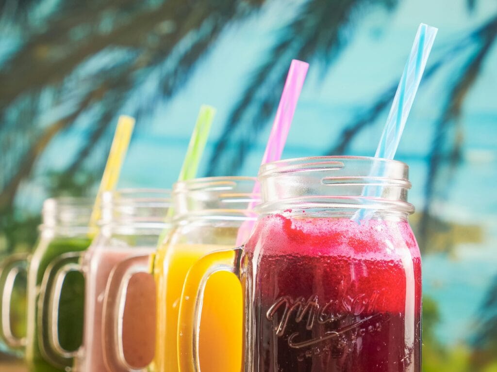 Colorful tropical juices in mason jars with straws against a blurred beach background.