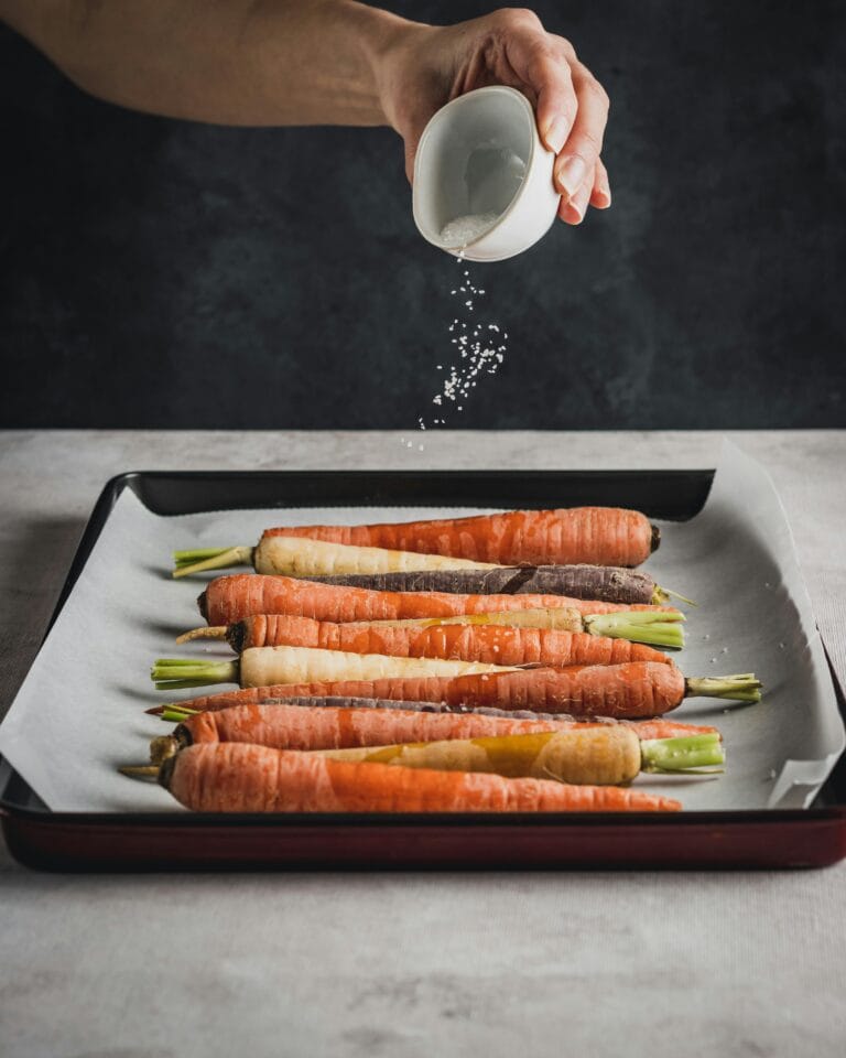 Hand sprinkling salt on vibrant carrots on a baking sheet, ready for roasting.