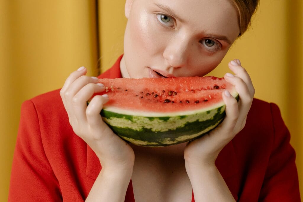 A young woman in a red jacket bites into a fresh watermelon slice against a yellow background.