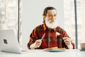 Senior man with beard enjoying pasta at home while working on a laptop. Capturing a cozy lifestyle moment.