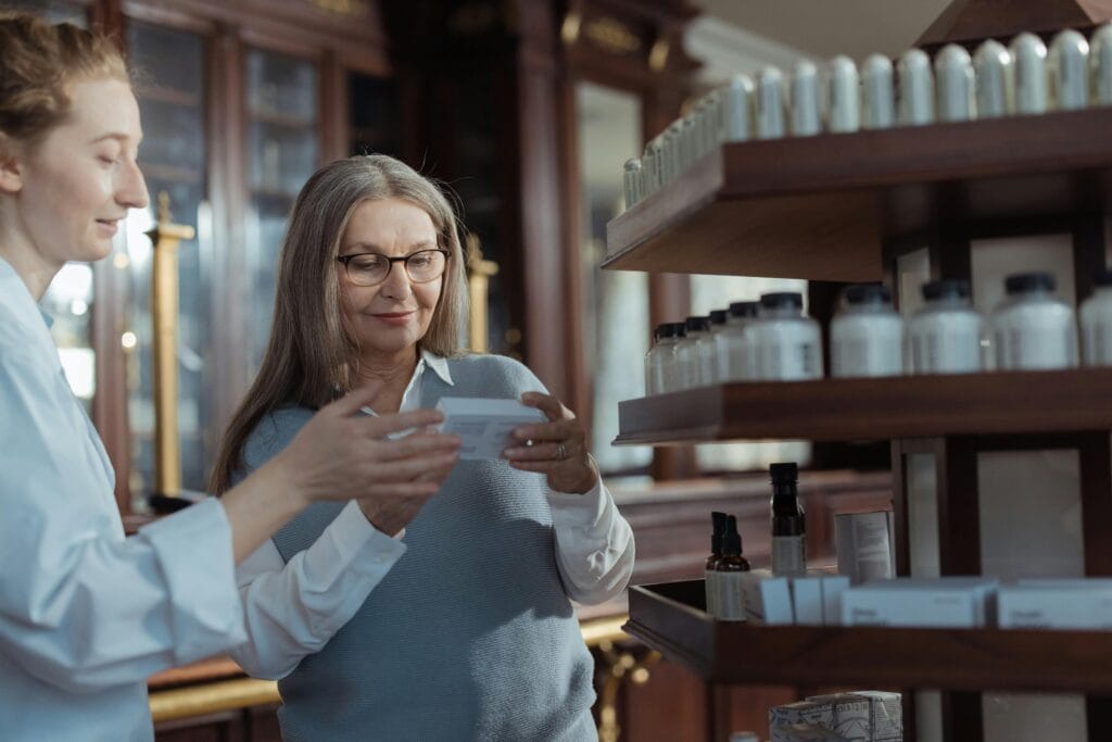 Elderly woman receiving assistance from a pharmacist in a pharmacy with product shelves.