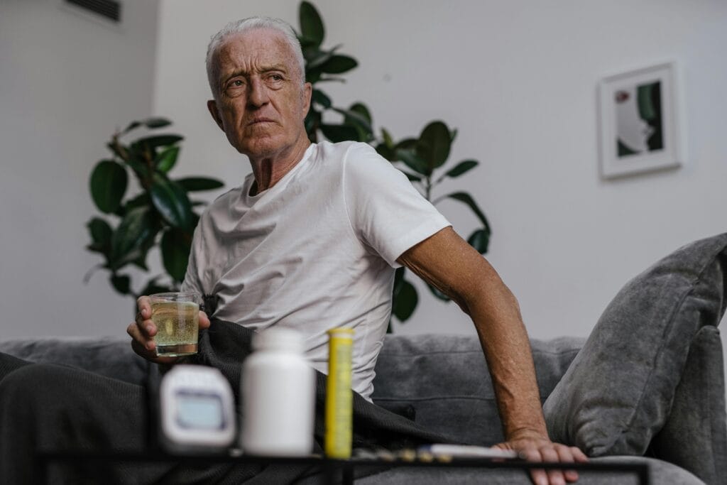 Senior man sitting on sofa with a drink, surrounded by plants and medication bottles.