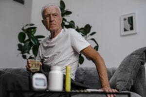 Senior man sitting on sofa with a drink, surrounded by plants and medication bottles.