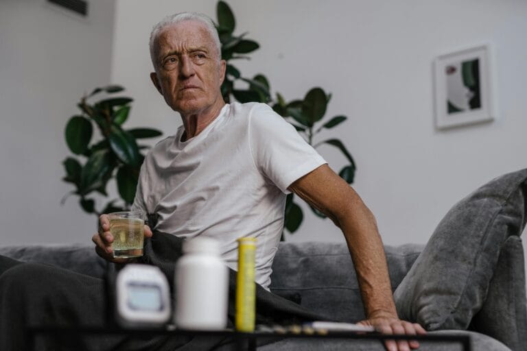 Senior man sitting on sofa with a drink, surrounded by plants and medication bottles.