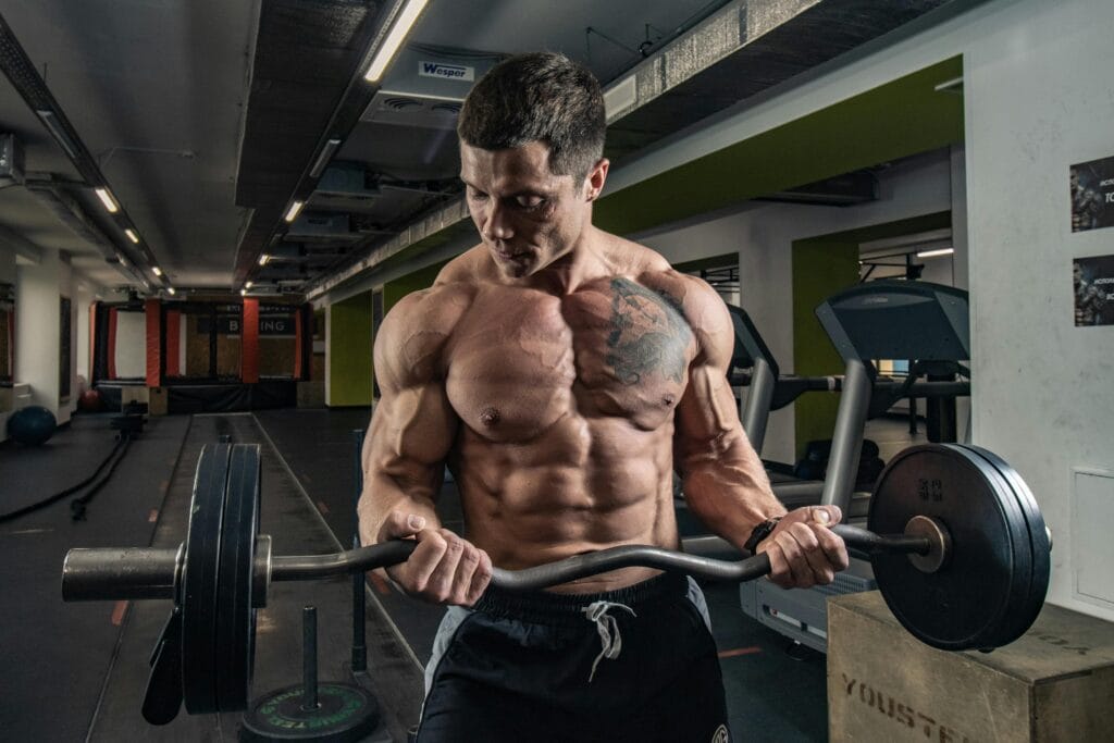 Topless bodybuilder with tattoos lifting weights in an indoor gym setting.