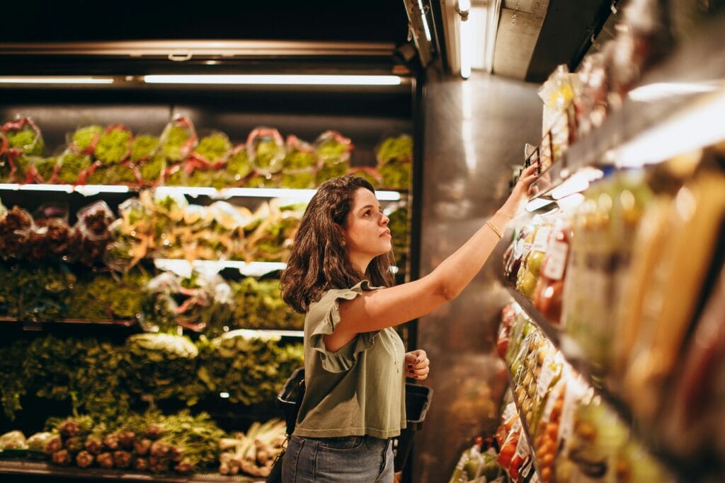 A woman selects fresh vegetables in a well-lit supermarket produce aisle.