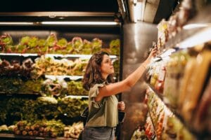 A woman selects fresh vegetables in a well-lit supermarket produce aisle.