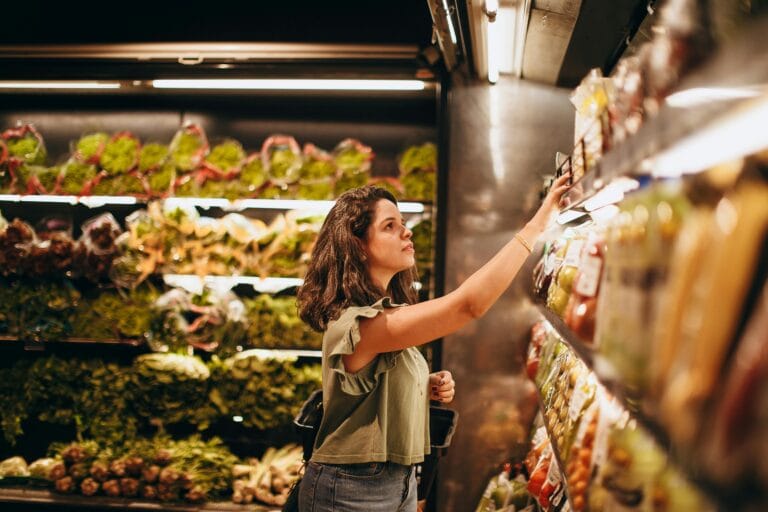 A woman selects fresh vegetables in a well-lit supermarket produce aisle.