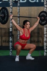 A female athlete wearing red activewear doing an overhead squat with a barbell indoors at a gym.