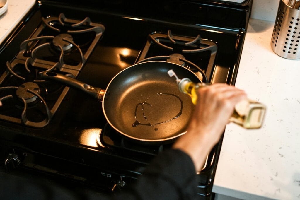From above crop faceless chef pouring olive oil into frying pan on stove while cooking at home