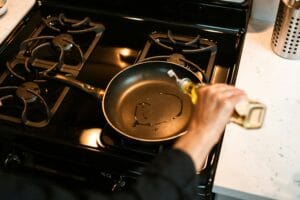 From above crop faceless chef pouring olive oil into frying pan on stove while cooking at home