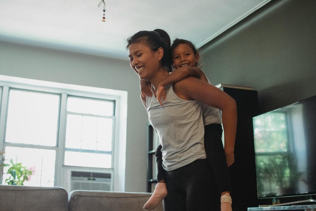 Laughing young female in casual clothes piggybacking adorable cheerful daughter while having fun together in modern living room