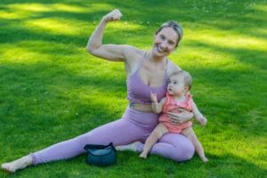 A smiling mother flexes playfully while sitting with her baby on a sunny lawn.