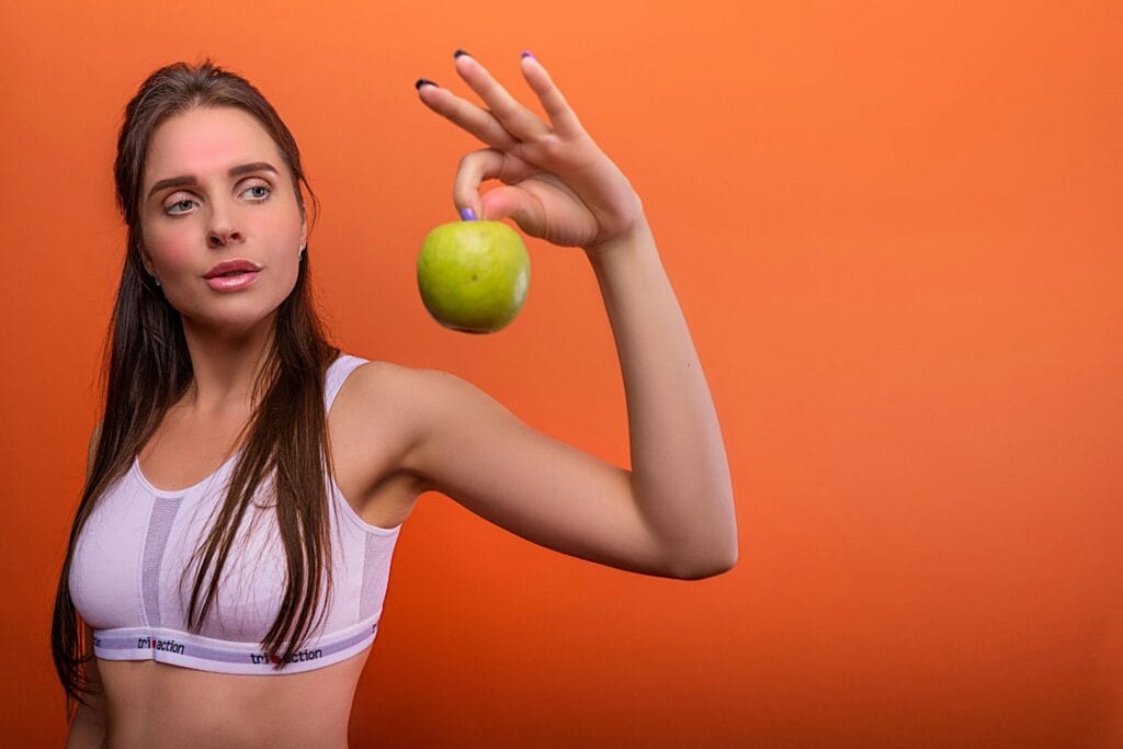 Young woman in activewear holding a green apple against orange background.
