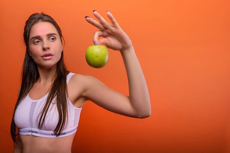Young woman in activewear holding a green apple against orange background.