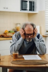 Senior man looking stressed while reading documents in a kitchen setting, suggesting financial or personal concerns.