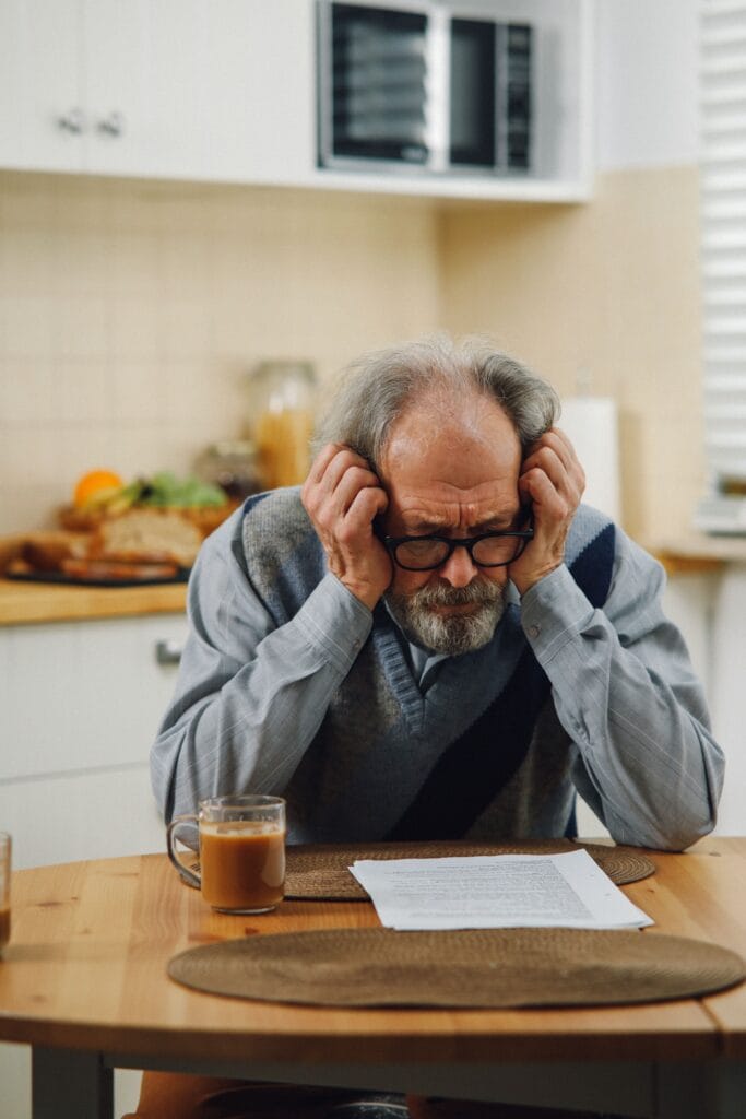Senior man looking stressed while reading documents in a kitchen setting, suggesting financial or personal concerns.