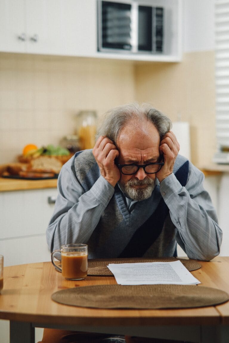 Senior man looking stressed while reading documents in a kitchen setting, suggesting financial or personal concerns.