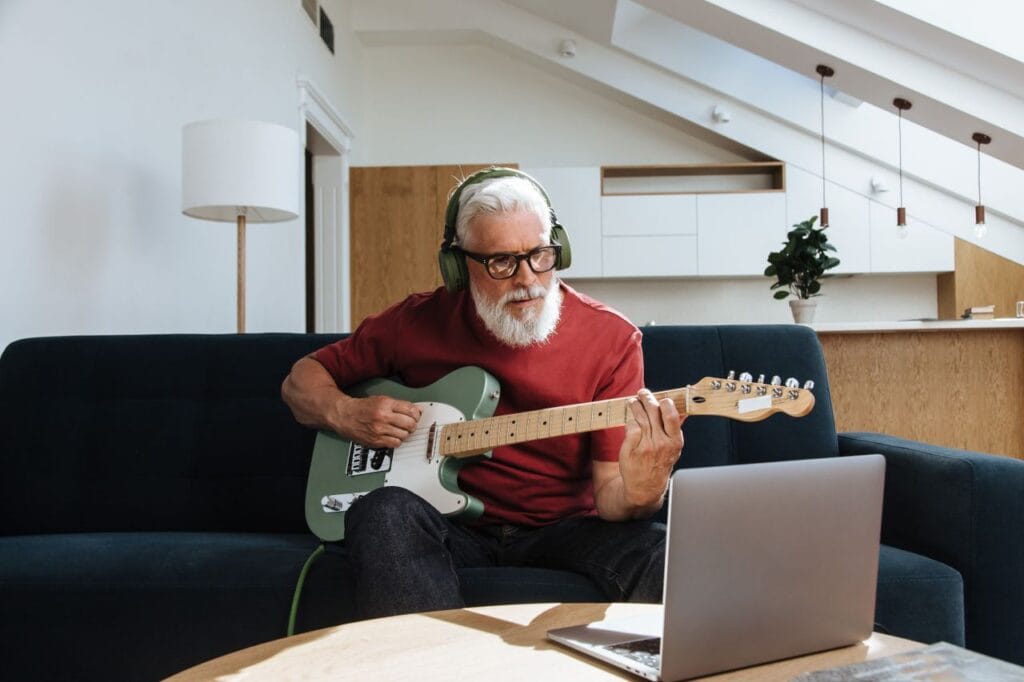 Elderly man enjoying music at home with electric guitar and laptop.