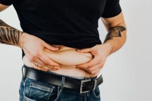 Close-up of a man pinching his stomach fold, wearing a black shirt and jeans with visible tattoos.