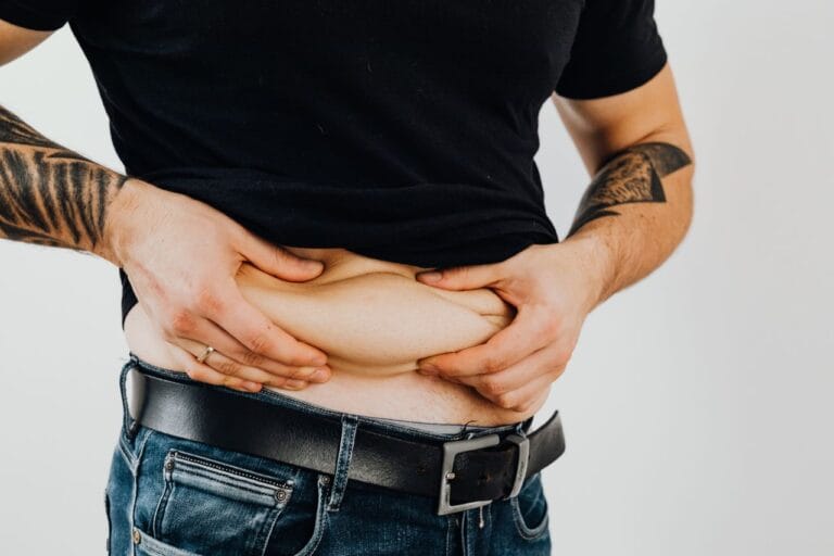 Close-up of a man pinching his stomach fold, wearing a black shirt and jeans with visible tattoos.