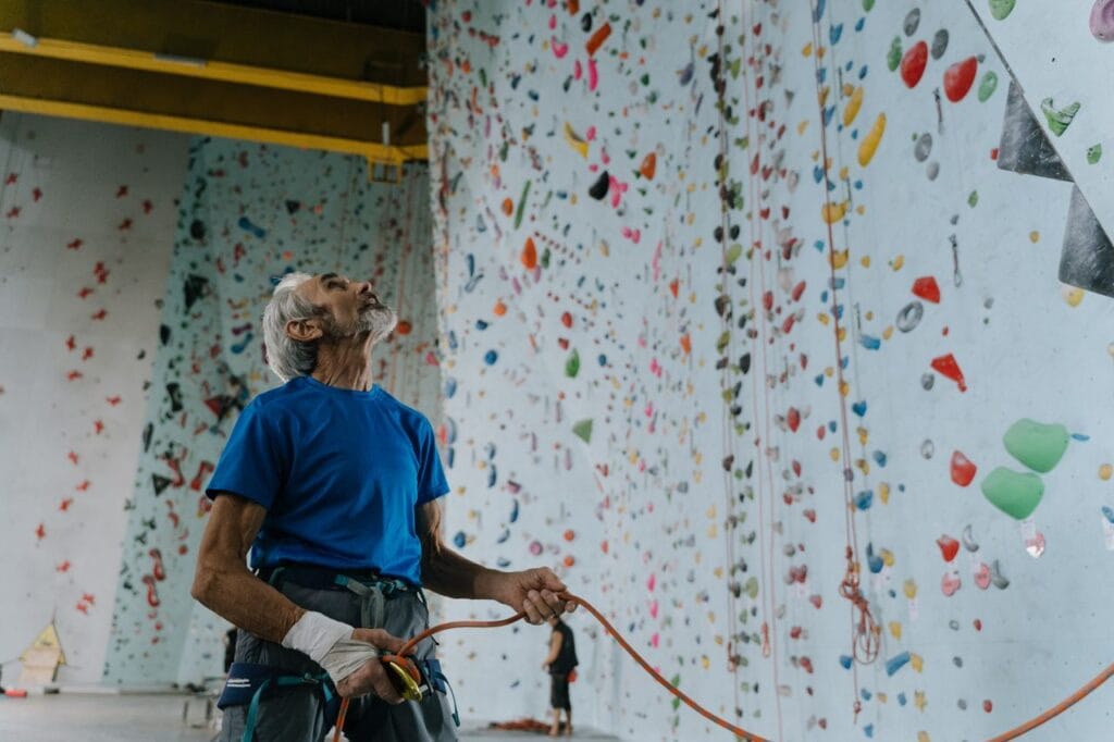 An elderly man in a harness preparing for rock climbing indoors, gazing at the wall.