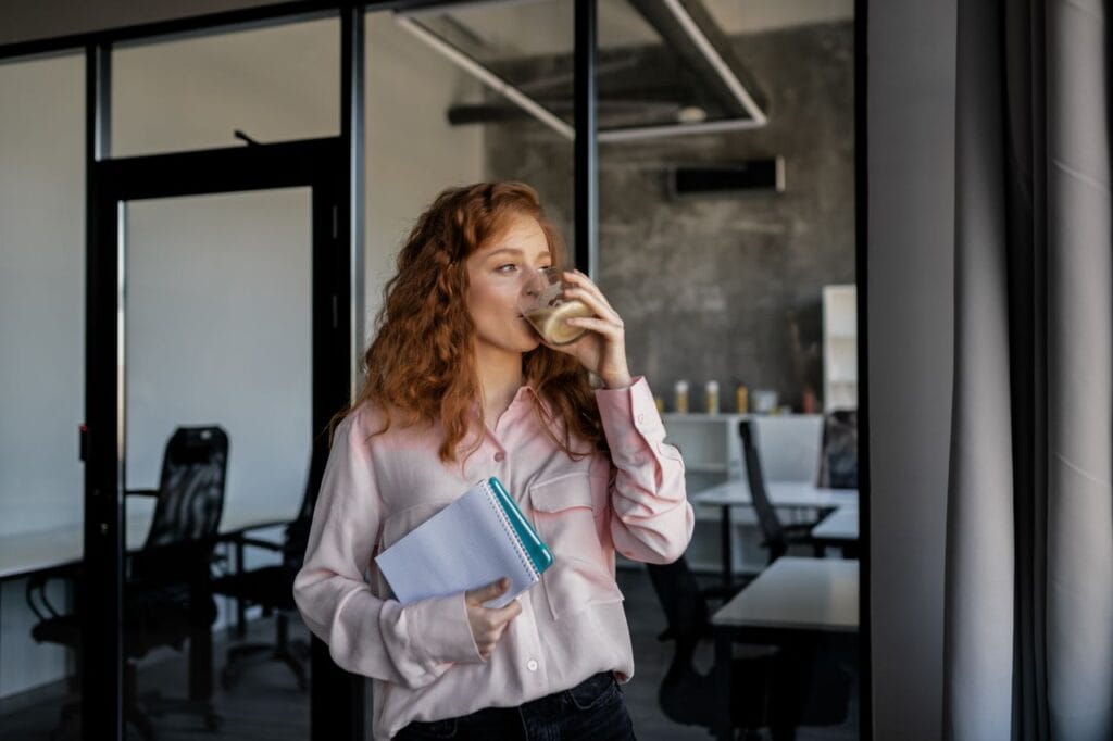 A redhead woman enjoys a coffee break while holding notebooks in a modern office.
