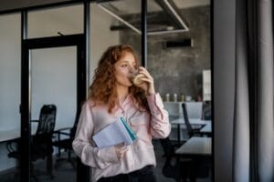 A redhead woman enjoys a coffee break while holding notebooks in a modern office.