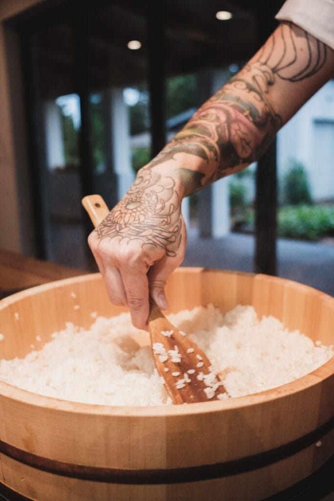 Close-up of a tattooed arm using a wooden spatula to stir rice in a kitchen setting.