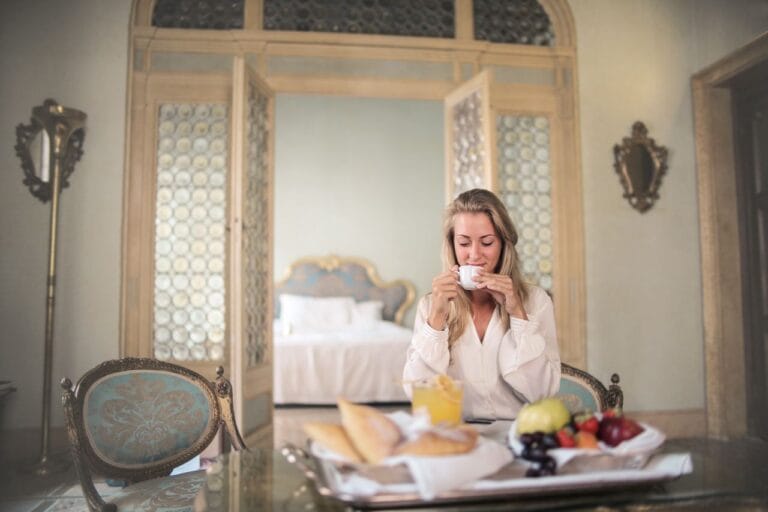 Glad female in elegant wear sitting at table with metal tray of healthy breakfast in vintage hotel room and drinking coffee in morning