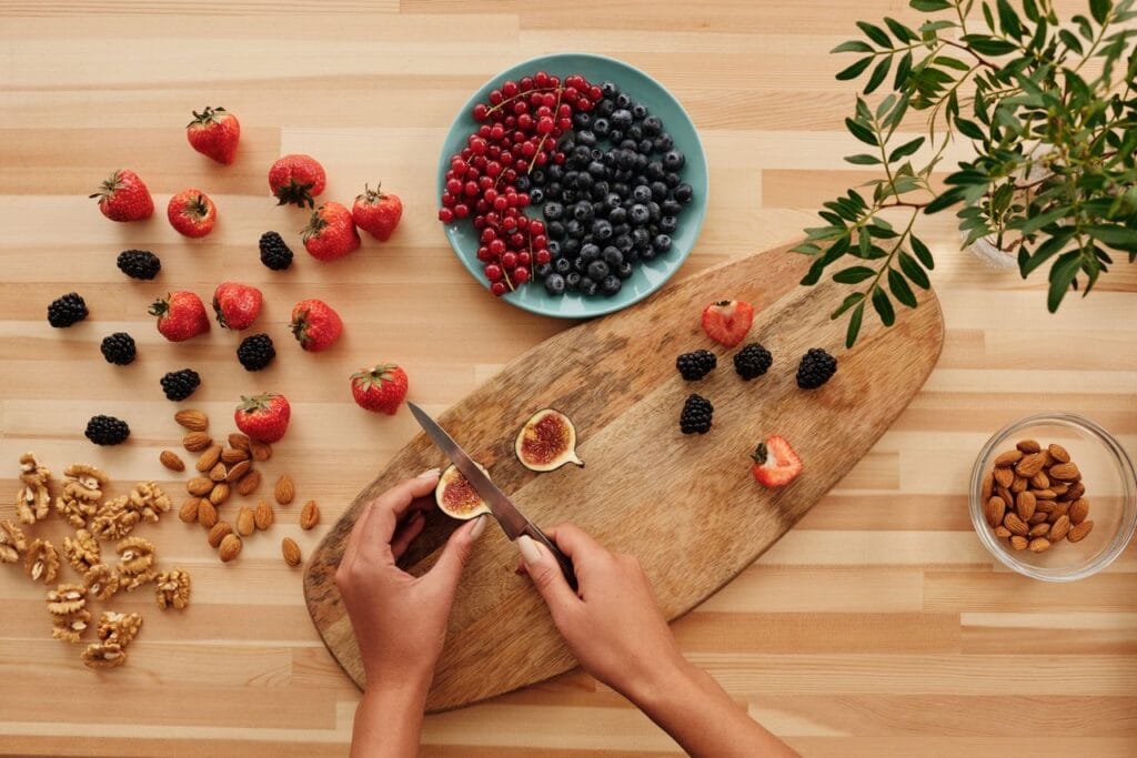 Overhead shot of fresh fruits, nuts, and hands preparing ingredients on a wooden board.