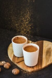 Two coffee cups on wooden heart-shaped board with cinnamon and nutmeg, against a dark background.