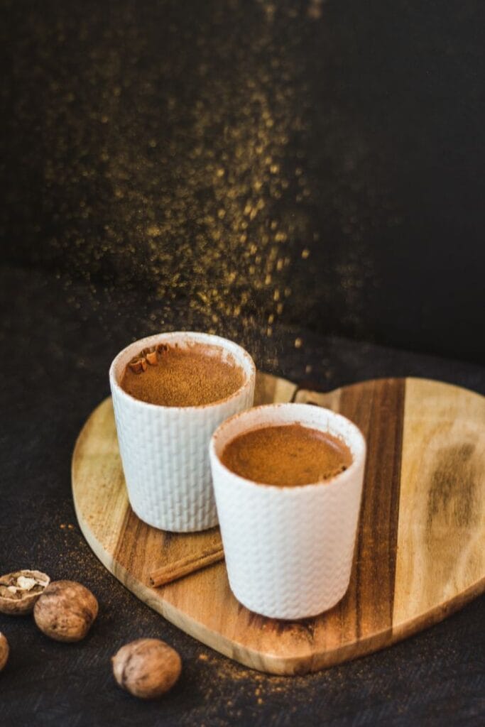 Two coffee cups on wooden heart-shaped board with cinnamon and nutmeg, against a dark background.
