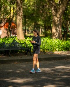 A woman in sportswear walks through a sunlit park while using her phone.