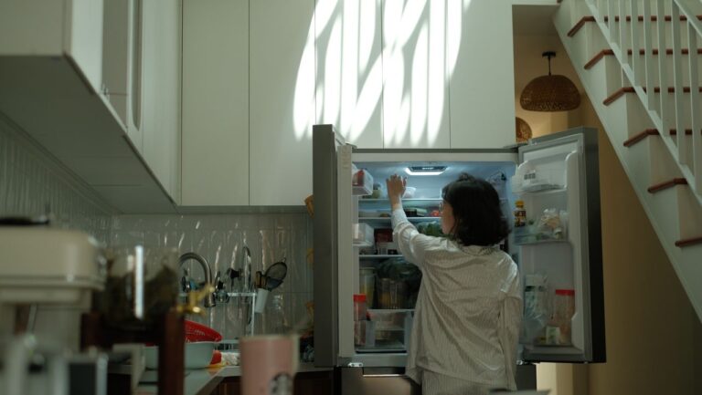 A woman in pajamas checks the refrigerator in a warmly lit cozy kitchen.